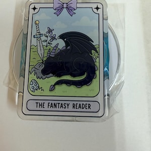 May include: A rectangular acrylic charm featuring a light blue background with a black dragon sleeping on green grass. A silver sword, purple flowers, and a spotted egg are visible. A purple bow sits at the top, and the text THE FANTASY READER is printed at the bottom.