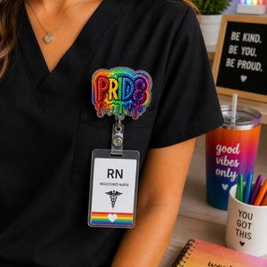 May include: A black scrub top with a colorful "PRIDE" badge and a "Registered Nurse" ID badge. The badge features rainbow colors and dripping paint effect. A rainbow-themed tumbler and notebook are also visible.