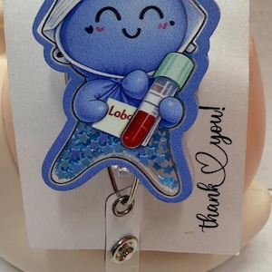 May include: A blue badge reel featuring a cartoon character holding a blood vial. The character is wearing a hat and has a smiling face. The badge reel has a clear plastic strap and the words "thank you!" are written on the attached card.