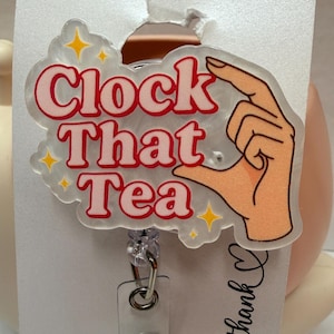 May include: A clear acrylic badge reel with the text "Clock That Tea" in red letters with a white outline. The design includes a hand gesture and yellow star accents. The reel has a clear plastic strap and a metal clip.