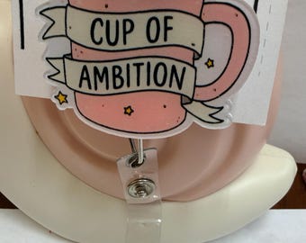 Könnte beinhalten: Ein rosa Ausweishalter mit einer Kaffeetassen-Illustration und einem Banner mit der Aufschrift CUP OF AMBITION. Das Design enthält kleine gelbe Sterne und ist an einem transparenten Kunststoffband mit einem Metalldruckknopf befestigt.
