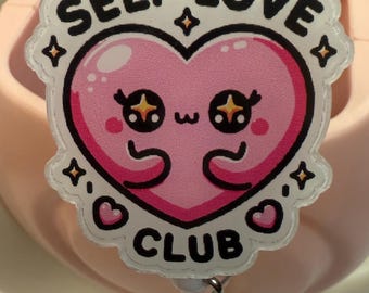 Könnte beinhalten: Weißer und rosa Selbstliebe-Club-Badge mit einem Cartoon-Herz mit funkelnden Augen und einem kleinen Herz auf jeder Seite. Der Badge hat einen silbernen Schlüsselanhänger.