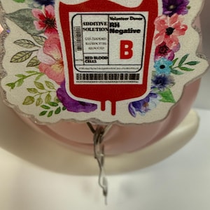May include: A red blood bag with a white label that reads "ADDITIVE RH SOLUTION Negative" and "B". The bag is surrounded by a floral wreath with pink, yellow, and purple flowers.