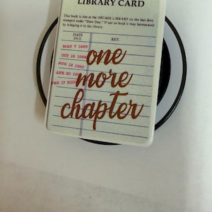 May include: A rectangular phone grip featuring a vintage library card design with blue lines and red text. The card reads LIBRARY CARD, followed by dates and the phrase one more chapter in large brown script. The grip is mounted on a black circular base.