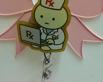 May include: A badge reel featuring a light green cartoon character wearing a white lab coat and a white cap with a red Rx symbol. The character is typing on a laptop with an Rx symbol on the screen. The design has a gold glitter border.