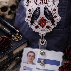 Pode incluir: Um crachá com o texto "F Caw F" e um desenho com dois corvos negros voltados um para o outro sobre um coração vermelho e rosas. O crachá está preso a um porta-cartão de plástico transparente com uma foto.