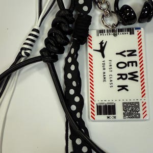 Puede incluir: Una cinta de lunares blancos y negros, un cordón de cuero negro y un cordón trenzado blanco y negro están unidos a un llavero plateado con un anillo en forma de corazón. Un amuleto de auriculares negro también está unido al llavero. Una tarjeta blanca con rayas rojas y el texto "NEW YORK FIRST CLASS YOUR NAME" está unida al llavero.
