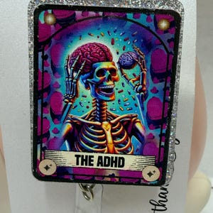 Könnte beinhalten: Ein quadratischer Ausweishalter mit einer farbenfrohen Grafik eines Skeletts, das ein Gehirn hält, vor einem Hintergrund aus schwebenden Pillen. Das Design enthält den Text THE ADHD in einem Banner unten, eingefasst in einen glitzernden Rand.