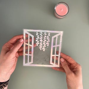Op de afbeelding: Een vierkante, iriserende raamsticker met een open raamontwerp en hangende bloemaccenten. De sticker wordt door handen vastgehouden, met een roze kaars in een glazen pot op de achtergrond.