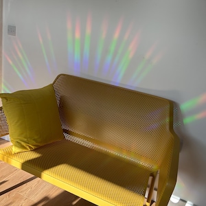 May include: A yellow metal bench with a perforated seat and backrest, paired with a matching yellow pillow. The bench is positioned against a white wall, with colorful light refractions illuminating the scene. The bench is on a wooden floor.