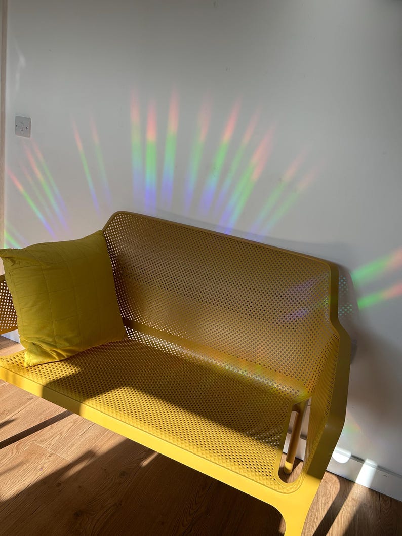 May include: A yellow metal bench with a perforated seat and backrest, paired with a matching yellow pillow. The bench is positioned against a white wall, with colorful light refractions illuminating the scene. The bench is on a wooden floor.