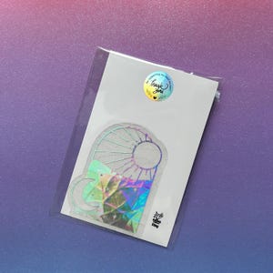 May include: A holographic sticker featuring a stained glass design with a sun, moon, and geometric shapes. The sticker is on a white card, sealed in a clear plastic sleeve. A round sticker with the words "Thank You" is on the top.