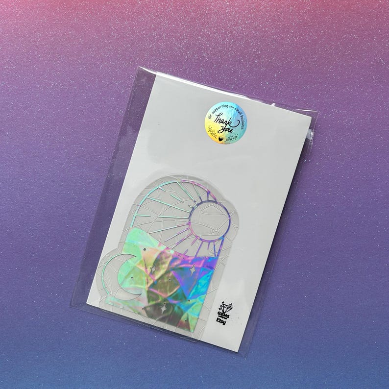 May include: A holographic sticker featuring a stained glass design with a sun, moon, and geometric shapes. The sticker is on a white card, sealed in a clear plastic sleeve. A round sticker with the words "Thank You" is on the top.