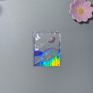 May include: A holographic sticker with a landscape design featuring a crescent moon and rolling hills. The sticker has a rainbow gradient effect and a clear border. A pink flower-shaped dish is in the background.
