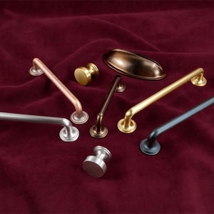 Puede incluir: Una colección de herrajes para armarios en varios acabados metálicos, incluyendo plata, cobre, oro y bronce. El surtido presenta pomos, tiradores y una concha, todo exhibido sobre una superficie de terciopelo rojo oscuro.