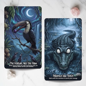 May include: Two rectangular oracle cards featuring dark, illustrative artwork. The left card shows a toucan with the text 'The Feature, Not the Flaw' and a subtitle. The right card depicts a crocodile in a swamp with the text 'Patience Has Teeth'.