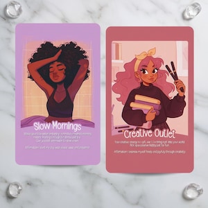 May include: Two illustrated affirmation cards. One card features a woman with dark hair, the text "Slow Mornings", and affirmations. The other card shows a woman with pink hair, the text "Creative Outlet", and affirmations. The cards are on a marble surface.