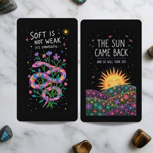 May include: Two rectangular black cards with colourful illustrations. The left card features a pink and purple snake among flowers with the text 'SOFT IS NOT WEAK IT'S STRATEGIC.' The right card shows a sun rising over floral hills with the text 'THE SUN CAME BACK AND SO WILL YOUR JOY.'