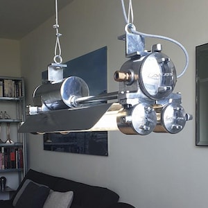 May include: A silver-toned industrial pendant light fixture suspended by two thin cables. The fixture features a cylindrical metal housing with multiple circular end caps, each embossed with the text "DAWSON". A horizontal light tube is visible.