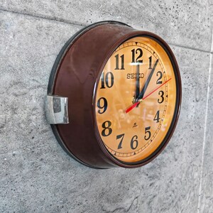 Peut inclure: Une horloge murale Seiko ronde, marron, avec un cadran orange clair. L'horloge présente des chiffres noirs, des aiguilles noires et une trotteuse rouge. L'horloge est fixée sur un mur gris texturé.