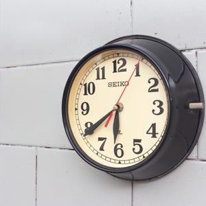 Könnte beinhalten: Eine schwarze Seiko Wanduhr mit cremefarbenem Zifferblatt und schwarzen Ziffern. Die Uhr hat einen roten Sekundenzeiger und ist an einer weißen gefliesten Wand befestigt. Die Uhrzeit ist ungefähr 6:40 Uhr.