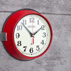 Op de afbeelding: Een ronde Seiko wandklok met een rode rand, witte wijzerplaat en zwarte cijfers. De klok heeft zwarte wijzers en een rode secondewijzer. De klok is bevestigd aan een grijze betonnen muur. De klok heeft een diameter van ongeveer 25 cm.