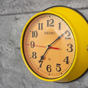 Puede incluir: Un reloj de pared Seiko amarillo con un borde negro y una esfera naranja claro. El reloj tiene números y manecillas negras, con un segundero rojo. El reloj está montado en una pared gris texturizada.