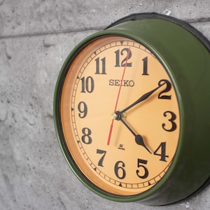 Può includere: Un orologio da parete Seiko vintage con cassa in metallo verde e quadrante color crema. L'orologio presenta numeri neri, lancette nere e una lancetta dei secondi rossa. Il logo Seiko è visibile nella parte superiore del quadrante.