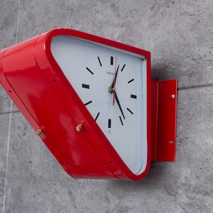 Puede incluir: Un reloj de pared rojo de estilo retro con esfera blanca y números negros. El reloj tiene un diseño curvo distintivo y está montado en una pared de hormigón gris. Las manecillas del reloj son negras y doradas. La marca "Seiko" es visible.