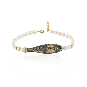 May include: A pearl bracelet featuring a decorative fish-shaped centerpiece. The fish is black with gold floral accents and small diamonds. The bracelet has a gold clasp and gold accents between the pearls.
