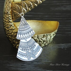 May include: Silver earrings with a geometric pattern. The earrings are in the shape of a triangle and have a layered design. The earrings are hanging from a gold-colored stand.