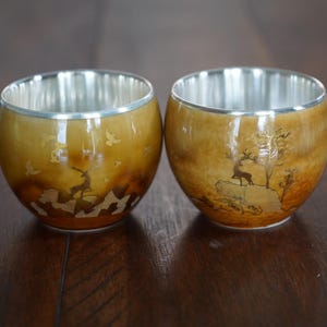 May include: Two silver-rimmed cups with brown and gold designs. The cups feature a deer and mountain scene.