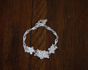 May include: A silver bracelet with a delicate floral design. The bracelet is made of filigree and features three flower motifs.