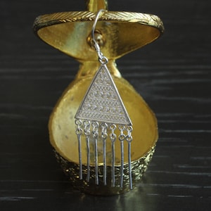 May include: A silver-toned earring with a triangular pendant. The pendant features intricate geometric patterns and dangling vertical bars. The earring is displayed against a gold-colored, ornate background, with a dark wooden surface.