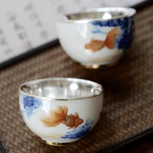 以下が含まれることがあります： 銀色の縁と金色のアクセントが付いた、白いセラミックティーカップが2つ。各カップには、青い水の中を泳ぐオレンジ色の金魚の詳細なイラストが描かれています。カップは、お茶やその他の飲み物用に設計されています。