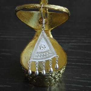 May include: Silver triangle earring with intricate filigree design and dangling teardrop-shaped beads. The earring is displayed on a gold stand.