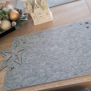Puede incluir: Un mantel individual de fieltro gris con recortes de estrellas. El mantel individual rectangular está sobre una mesa de madera. Hay adornos navideños en el fondo.