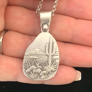 May include: Silver pendant necklace featuring a desert landscape design. The pendant showcases a detailed relief of a saguaro cactus, mountains, and desert plants. The necklace is held by a hand.