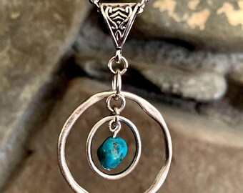 May include: A silver necklace with a turquoise stone pendant. The pendant is a double circle design with a smaller circle inside the larger circle. The turquoise stone is in the center of the smaller circle.
