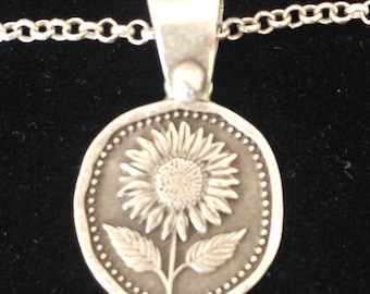 Könnte beinhalten: Silberne Anhänger Halskette mit einem Sonnenblumen-Design. Der runde Anhänger zeigt ein erhabenes Sonnenblumen-Bild und einen gepunkteten Rand. Die Kette ist im zarten Gliederstil gehalten, passend für den täglichen Gebrauch.