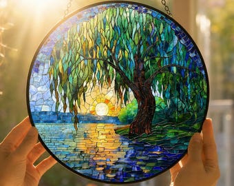 May include: A circular stained glass suncatcher featuring a mosaic design of a willow tree by a lake at sunset. The scene uses shades of blue, green, and yellow glass pieces to depict the tree, water, and sun, all encased in a black metal frame.