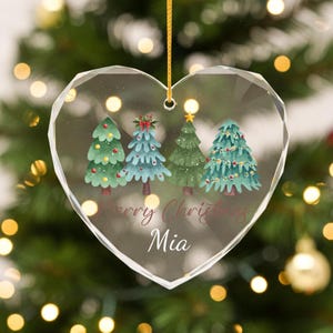 May include: Heart-shaped Christmas ornament featuring four watercolour Christmas trees in shades of green and blue. The ornament has the words "Merry Christmas Mia" in a cursive font and hangs from a gold string.