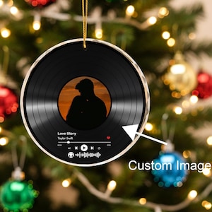 May include: A round, black Christmas ornament with a vinyl record design. The center features a silhouette of a couple kissing against an orange sunset. The text "Love Story" and "Taylor Swift" are visible, along with a music player interface.