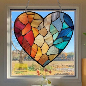 May include: A stained glass heart suncatcher with a black border, suspended by a chain. The heart is composed of colorful, irregular glass pieces in shades of red, orange, yellow, blue, and green. The suncatcher hangs in front of a window.