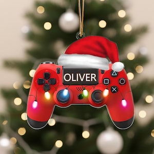 May include: A red video game controller Christmas ornament with the name "OLIVER" on it. The ornament is decorated with a Santa hat and small lights. The ornament is hanging in front of a Christmas tree.