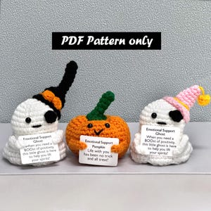 May include: Three handmade crochet Halloween decorations: two ghosts and a pumpkin. The ghosts are white with black accents and hats. The pumpkin is orange with a green stem. Each holds a sign with text.