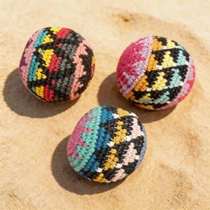 Puede incluir: Tres bolas hechas a mano con ganchillo y patrones geométricos en tonos rosa, azul, amarillo, negro y blanco. Las bolas se exhiben sobre arena, mostrando sus colores vibrantes y texturas.
