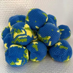 May include: A pile of blue, yellow, and green crocheted Earth globes. The globes are handmade with blue representing the oceans, yellow for continents, and green for landmasses. The handmade spheres are clustered together.