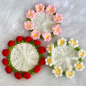 May include: Three crocheted coasters with floral designs. The first coaster features red roses, the second features pink flowers, and the third features white daisies.
