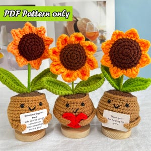 以下が含まれることがあります： 笑顔の顔が付いた茶色の鉢に入った、3つの編みぐるみのヒマワリの植物。ヒマワリはオレンジ色の花びらと茶色の中心部を持っています。1つの鉢には赤いハート、別の鉢には「成長を手伝ってくれてありがとう」と書かれたサイン、3つ目には「Emotional Support Plant」と書かれたサインが入っています。